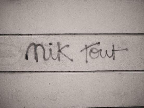 L’image montre un graffiti noir écrit sur un mur beige, avec l’inscription “Nik tout”.