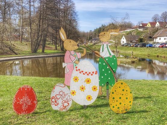 Osterdekoration vor dem Schloss Vildštejn in Skalná, Tschechien. Ein weiblicher und ein männlicher Osterhase und vier farbige Ostereier vor der Kulisse eines Teiches.