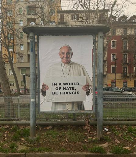 Poster-style street art by Andrea Villa displayed in a glass-covered frame on a sidewalk in an urban area. The artwork features a smiling Pope Francis holding a sign that reads: “IN A WORLD OF HATE, BE FRANCIS.” Leafless trees and apartment buildings are visible in the background on a cloudy day in what appears to be an Italian city.
