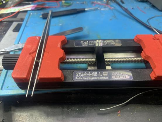 A double ended PCB vise, holding a pair of tweezers in one end.  The tweezer tips protrude from the side of the vise.