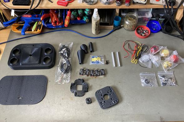 Parts for the Open Source Arcade Stick spread out on my workbench. The case parts are 3d printed, there are some buttons and microswitches, a PCB for auto fire and various small bits and pieces.