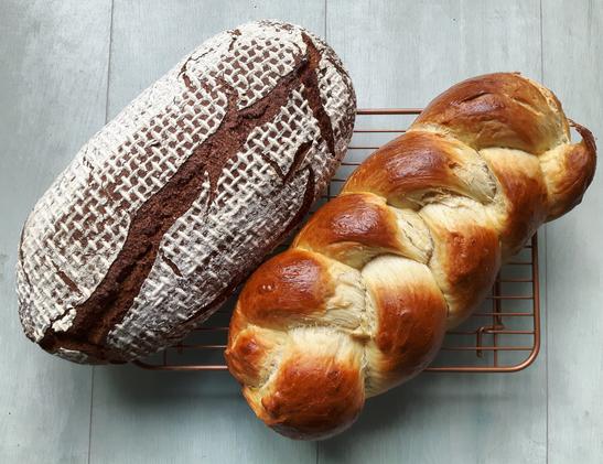 Ein dunkelbrauner bemehlter Brotlaib und und goldbrauner Hefezopf nebeneinander auf einem Abkühlgitter.