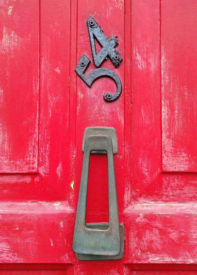 a close-up on a part of a wooden red door with a metal piece to knock on the door and a black metallic 5 and 4 nailed in a very crooked way to fit the center frame piece of the door.
