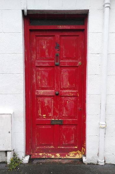 A wooden red door with peeling paint, we can start seeing some yellow under it in places. in the middle highest part of the door, the "54" number is nailed crooked to fit the frame of the door