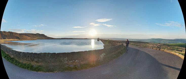Reservoir on a hillside, more hills beyond.  Sun low on the horizon, reflected in the water.  Small dog hidden in the shadow of the reservoir's low wall