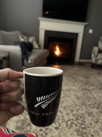 A black and white mug with the word, “New Zealand” on it being held in front of a cozy fire on a quiet living room.