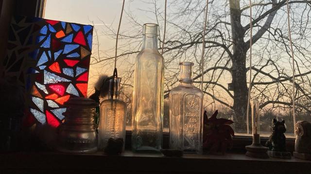 Objects on s windowsill are catching the day’s first light. Left to right: A “stained glass” school art project, three antique bottles, small knick knacks.  Outside the window the trunk and bare branches of a sycamore tree. Pastel orange and blue sky.