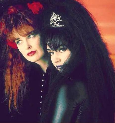 Jill Bryson with red crimped haid, looking away from camera. Immediately in front of her is Rose McDowall in a black leather catsuit, with black crimped hair and a tiara, looking at the camera.