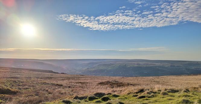 Moorland falling off into valley with a village spread out below.  Blue sky above the flat horizon of the other side of valley, with a low Sun and a patch of scaley, white cloud