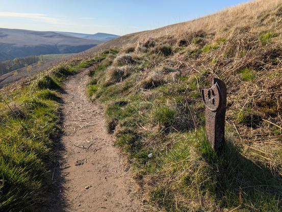 Pitted iron post, proclaiming a capital 'G', standing next to a footpath running along a moorland valley.  Beyond the curving closer horizon, a salience (Pule Hill) pokes out above the far horizon