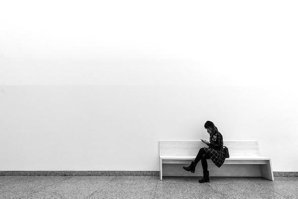 Black & white photo of woman in plaid coat and black boots sitting on a white bench in front of a white wall, scrolling her phone.