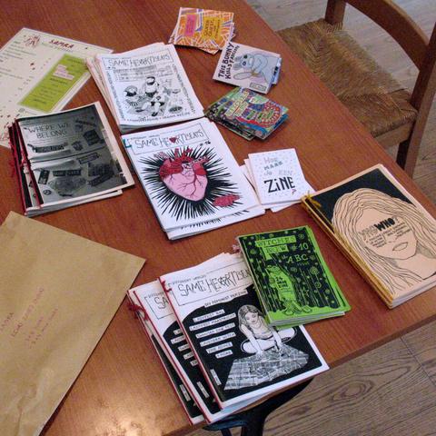 Zines, menu, and big envelope spread over the table. Zine titles include Same Heartbeats, Queer Quest, YngWHO, This Bunny kills fascists, and Witches' Brew.
