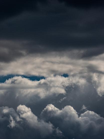 Aerial view of a dramatic cloudy sky. The image shows multiple layers of clouds with varying textures and densities. The top portion features dark, heavy storm clouds in deep blue-gray tones. The middle section reveals a break with some patches of blue sky visible between cloud formations. The lower portion displays fluffy cumulus clouds with white and light gray coloring. The overall scene captures the dimensional nature of cloud formations at different altitudes, creating distinct layers throughout the atmosphere.