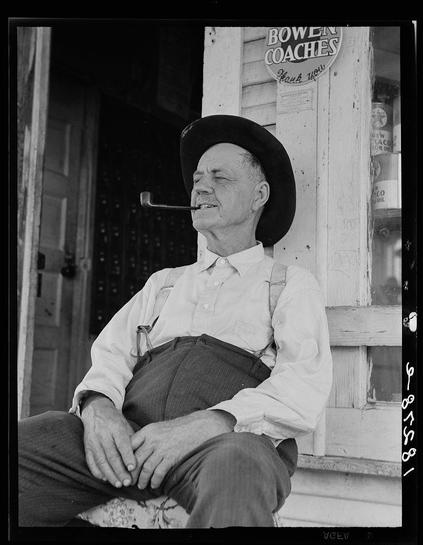 This black and white photograph features an elderly man sitting in front of a wooden structure. He is wearing suspenders, rolled-up sleeves on his shirt, and appears to be smoking a pipe while looking off into the distance with a thoughtful expression. His left hand rests gently across his lap, holding what might be a floral-patterned cloth or sleeve protector.
Behind him, there's signage that reads "BOWEN COACHES," suggesting he may be associated with a coach company named Bowen Coaches or perhaps in front of one of their facilities. The image is marked by the number 68-07251 and has been processed as per GOCV standards.
The overall atmosphere evokes a sense of nostalgia, reflecting on past times where individuals like O.O. Mills struggled to make ends meet amidst changing agricultural technologies such as tractors that were hindering their livelihoods in Childress County, Texas during the time captured by Dorothea Lange's photography work.