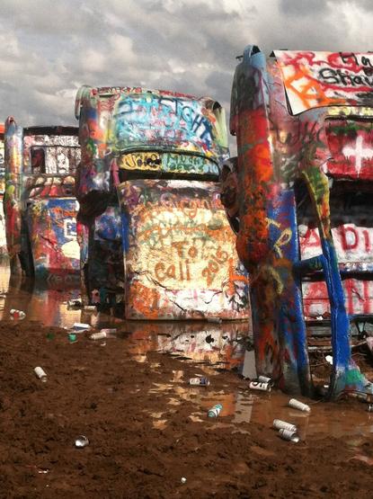The once-famous Cadillac Ranch art installation with the classic vehicles sticking out of the ground, but now littered with leftover spray paint cans, fully covered in bad graffiti, and surrounded by deep puddles of mud.