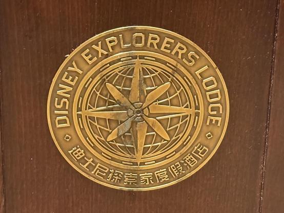 A bronze medallion featuring the text "DISNEY EXPLORERS LODGE" surrounding a compass and globe design. The background is wood.