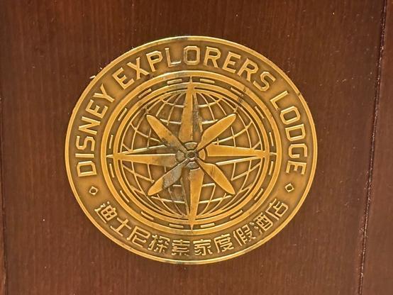 A bronze medallion featuring the text "DISNEY EXPLORERS LODGE" surrounding a compass and globe design. The background is wood.