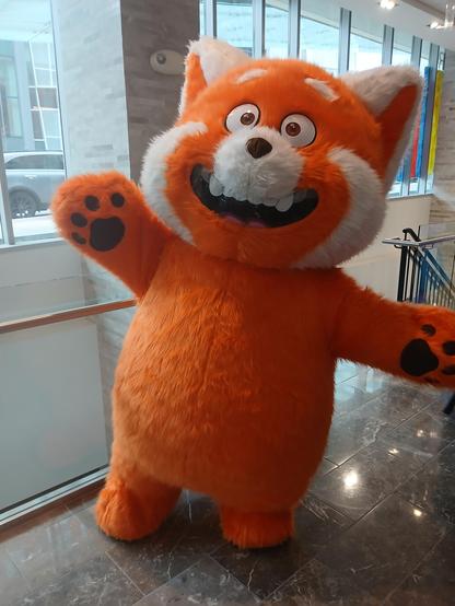 A person wearing a red panda fursuit costume of the red panda from Turning Red. It looks exactly like in the movie. They are striking a happy pose.