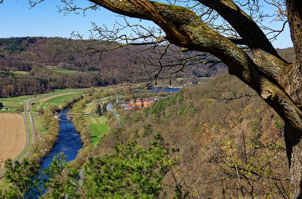 Blick auf einen Fluss, der sich in einem grünen Tal schlängelt. Ganz im Vordergrund rechts ragen Äste ins Bild. An der rechten Flussseite  ein klener Ort und eine Straße, links eine Eisenbahnlinie.