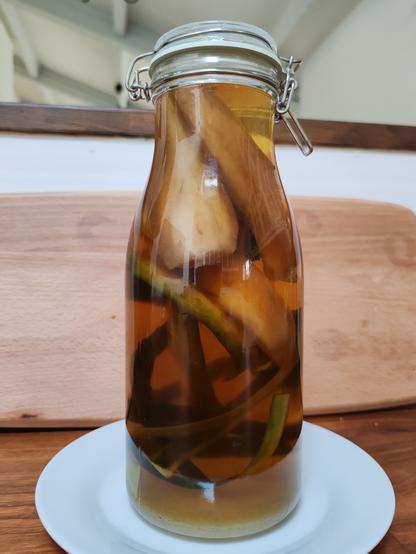 Tall glass jar in the shape of a wide necked bottle, with a kilner style closure, on a small white plate. The bottle contains a clear brown liquid, and has pieces of peeled pear and cucumber skins floating in it.