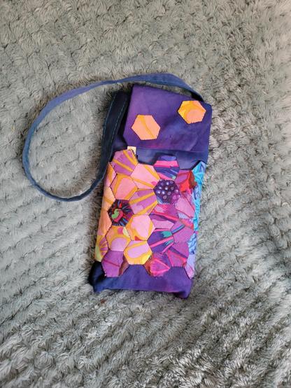 Photograph of the front of the power bank case. The case is a little rectangular bag with a strap for easy carrying and a flap over the top which closes with a hidden snap. It's made of purple and blue tie dye cotton left over from making a summer dress and has a pocket running all the way around the outside made of a patchwork of half inch hexagons. Those are made from tiny scraps of Kaffe Fasset fabric left over from making a quilt. The small hexagons are in shades of yellow, peach, purple, pink and blue with accents of light green and red.