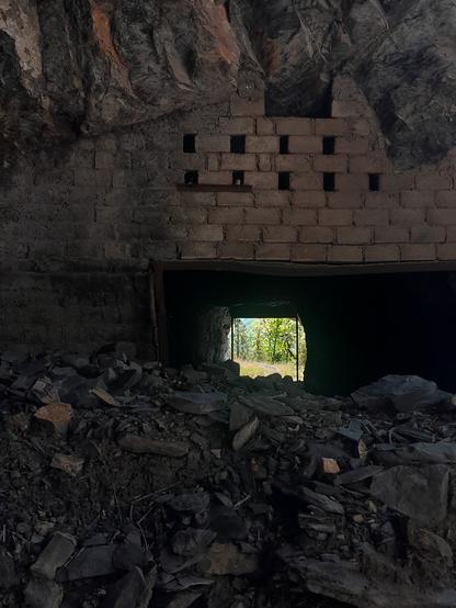 Blick in einen kurzen Tunel. Der Tunneleingang ist Gemauert. Er ist bis zur Hälfte mit Steinen und Erde verschüttet. Aus dem Tunel sieht mensch ins helle, sonnige Frühlingsgrün
