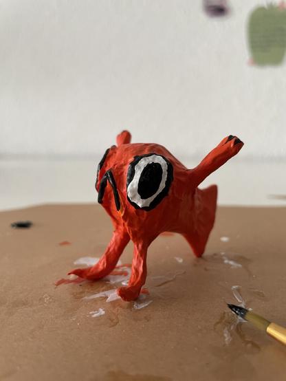 Kleine Pappmaché Figur leicht schräg fotografiert: orangener Fisch mit armen und Beinen auf einem Stück Pappe mit Farbklecksen vorn rechts im Bild ein Pinsel