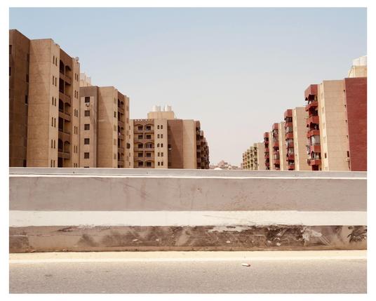 This image shows a view of multiple residential buildings, probably in an urban settlement. These buildings, appearing in shades of beige and red, have modern architectural features and several balconies visible on their facades. In the foreground, there is a low concrete barrier or wall, likely part of a road infrastructure, showing some signs of wear and staining. The sky is almost clear, indicating a sunny day. The general atmosphere suggests a warm and arid climate, possibly characteristic of regions like North Africa or the Middle East.