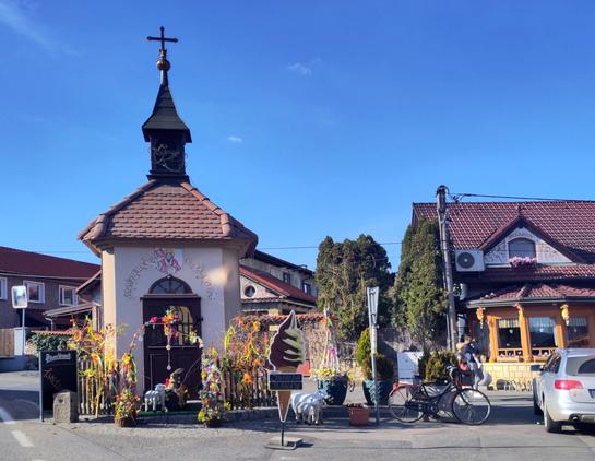 Small square chapel with red tiled roof and a dark bell tower topped by a cross. The wall is painted with a crest of a king with an orb and sceptre and the czech arms. There are lots of flowery decoration arounds among witch a big ice cream add. Drooderfiets stands on the right.