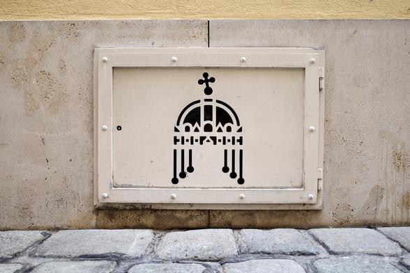 The image shows a small, beige metal window set into a stone wall just above a cobblestone street. On the door is a black stencil-like symbol resembling a stylized crown with hanging ornaments and a cross on top.