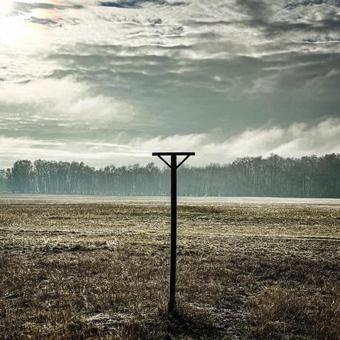 A solitary wooden post stands like a silent sentinel in a misty field under a  turbulent - cloud‑laden sky. The wide‑open landscape and muted tones evoke the raw intensity of an Amenra album cover - conjuring themes of solitude and existential longing. (Hamburg, Boberger Dünen)