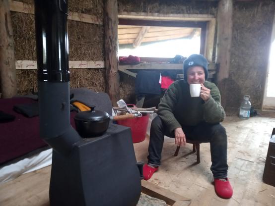 A woman sitting on a three legged stool in front of a small wood stove. Behind her, the walls between the pillars are still unrendered straw.

A cast iron pot sits on the stove.