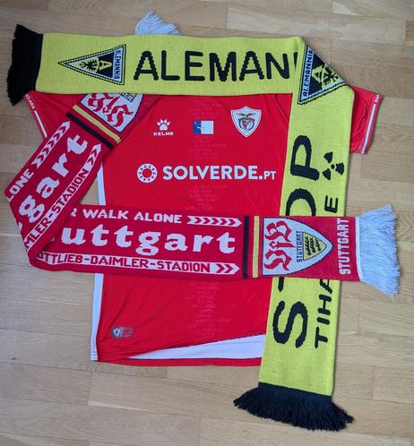 Ein rotes Fußballtrikot des portugiesischen Vereins CD Santa Clara liegt flach auf einem Holzfußboden. Auf dem Trikot sind die Umrisse der Azoren-Inseln sowie das Vereinslogo und mehrere Sponsoren zu sehen. Um das Trikot sind zwei Fanschals drapiert: Ein gelb-schwarzer Schal von Alemannia Aachen mit dem Vereinsnamen und dem Logo, sowie ein rot-weißer Schal des VfB Stuttgart mit der Aufschrift „You’ll never walk alone“. Der Schal zeigt ebenfalls das Vereinswappen des VfB Stuttgart.