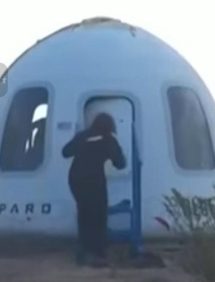 A woman in a blue jumpsuit leaning towards the door that is open slightly.