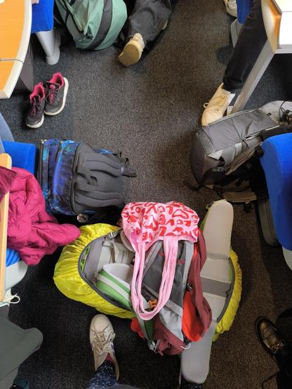 Das Bild zeigt eine Ansammlung von Rucksäcken und Taschen auf dem Boden eines Zugabteils. Im Hintergrund sind die Füße und Teile der Kleidung von Personen zu sehen, die auf den Sitzplätzen sitzen.
