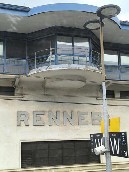 Das Bild zeigt die Fassade eines Gebäudes am Bahnhof mit der großen Aufschrift "RENNES" in großen, silbernen Buchstaben. Das Gebäude hat eine moderne Architektur mit großen Fenstern und einem markanten, geschwungenen Balkon im oberen Stockwerk, der mit einem blauen Rahmen versehen ist. Die Fassade ist in einem hellen Beige gehalten und weist einige Gebrauchsspuren auf.
