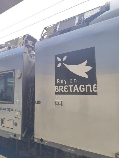 Das Bild zeigt einen Teil eines Zug, der an einem Bahnsteig steht. Auf der Seite des Zuges ist ein großes, dunkelblaues Schild mit einem weißen Symbol zu sehen, das eine stilisierte Vogel- oder Blattform darstellt. Unter dem Symbol steht in weißer Schrift "Région BRETAGNE".