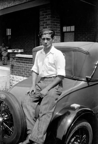 A man sits on the trunk of an old (not old at the time) Ford car that's parked outside a brick house. A man and a young child can be seen on the porch of the house.