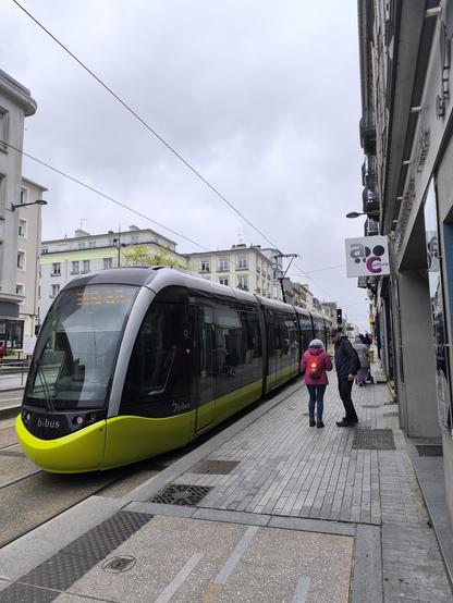 Eine Straßenbahn an der Haltestelle. Die Straßenbahn ist modern und Limetten-grün. Rechts im Bild ist ein Fußgängerweg und die Hauswand zu sehen.