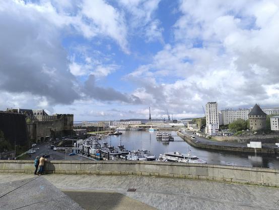 Blick auf den Militärhafen von Brest. In der Bildmitte sind einige Militärboote zu sehen, links Teile der Burg von Brest, rechts der Tour Tanguy. Der Himmel darüber ist bewölkt, aber es gibt ein paar blaue Stellen.