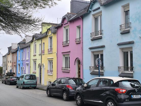 Das Bild zeigt eine Reihe farbenfroher Häuser. Die Häuser sind in Pastellfarben gestrichen, darunter Blau, Lila, Gelb und Rosa, und haben schräge Dächer mit dunkelgrauen Ziegeln. Jedes Haus hat mehrere Fenster mit weißen Rahmen und kleine Balkone mit Metallgeländern. Die Häuser sind in einer Reihe angeordnet und haben eine einheitliche Architektur mit weißen Türen und Fensterläden. Vor den Häusern sind mehrere Autos geparkt.