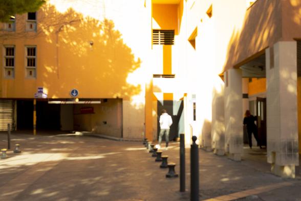 The image shows an urban scene with a yellow building. There are some architectural elements such as windows and columns, as well as shadows cast by trees or other structures. Two people are visible, one on the left next to a wall and another entering or exiting a doorway on the right. The setting appears sunny and possibly late afternoon given the long shadows. The area resembles a walkway or courtyard.