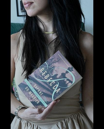 Woman in beige dress holds up Rebecca by Daphne du Maurier