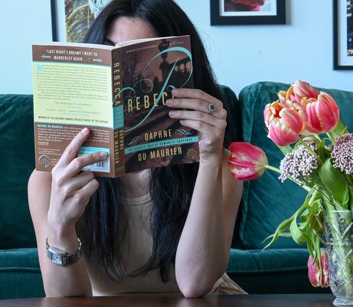 Woman, face covered, reads Rebecca by Daphne du Maurier. Flowers next to her.