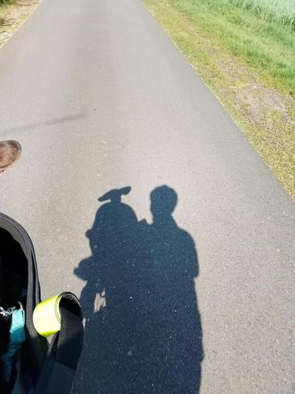 Schattenspielerei - wir haben die Sonne im Rücken und sie zeichnet unsere Silhouetten auf den Asphalt einer schmalen Straße. Effies Ohren wehen im Wind. Dahinter ist mein Umriss erkennbar. Ganz am Rand ist noch ein kleines Stück vom Fahrradkorb und ein Ohr von Effie zu sehen.