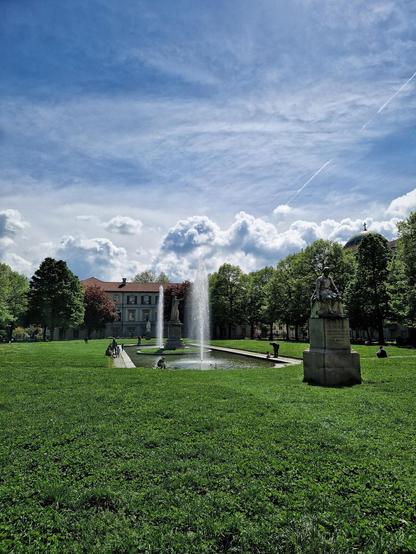 The image shows a grassy park area with a prominent fountain in the center. There are statues around the fountain, and several people are sitting on the grass and along the fountain's edge. In the background, there's a building surrounded by trees, and the sky above is mostly clear with some clouds.