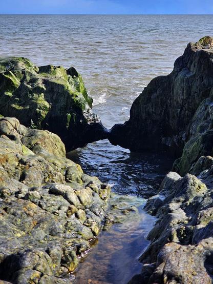 Water flows between moss-covered rocks towards the open sea under a partly cloudy sky. Two of the rocks are joined in an arch above the inflowing water below.