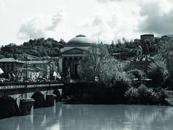 This is a black and white image depicting a scenic view of a river with a bridge spanning it. In the background, there's a prominent building with a dome and classical architecture, likely a temple or significant historical structure. The surrounding area features lush trees and hillside architecture, contributing to a picturesque, serene atmosphere.