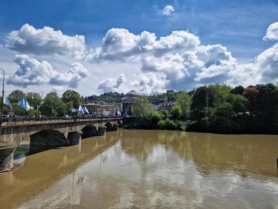 The image shows a scenic view of a river with a bridge crossing over it. The bridge has several arches and is lined with flags. On the far side of the river, there is a large, dome-shaped building, partially obscured by trees. The sky is filled with fluffy clouds, and there are hills in the background. The overall scene is peaceful and picturesque.