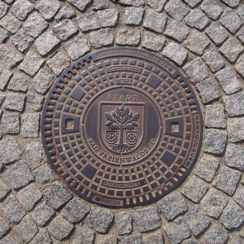 Round, metal manhole cover in the middle of cobblestones. It shows the lettering “Stadt Bad Freienwalde /O” and the town's coat of arms with an oak tree and two six-spoked wheels.